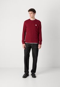 Bordeaux langemouwen overhemd met ribbelstructuur en een klein logo, gecombineerd met donkere, rechte jeans en zwarte schoenen. Minimalistisch ontwerp.
