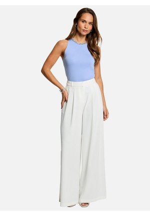 Femme portant un haut sans manches bleu clair avec un col orné de perles et un pantalon blanc à taille haute et jambes larges, debout avec une main dans la poche.