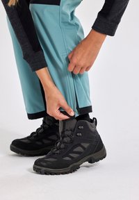 Person adjusting the cuff zipper on turquoise outdoor pants, wearing black and gray hiking boots on a white background.