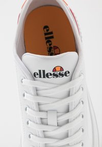 Sneaker in pelle bianca con superficie testurizzata, punta tonda e suola in gomma. Presenta lacci bianchi e un logo arancione e rosso all'interno.