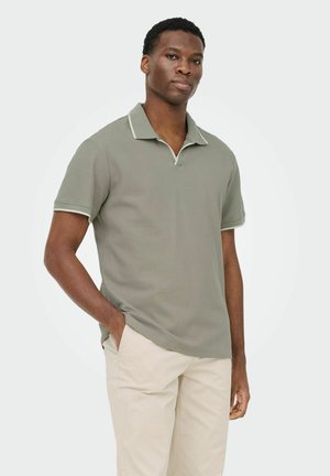 Mann trägt hellgraues Polohemd mit kurzen Ärmeln und beigefarbene Hose, steht mit einer Hand in der Tasche vor schlichtem Hintergrund.