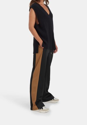 Personne portant un haut noir en maille sans manches avec un col en V et un pantalon noir ample avec de larges bandes latérales marron, debout avec une main dans la poche.