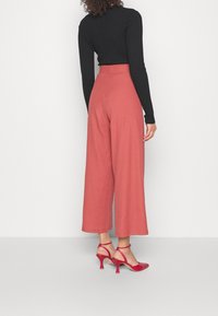 Brede broek in zacht koraalstof, hoge taille, gecombineerd met rode hoge hakken, met een getextureerde zwarte aansluitende top.