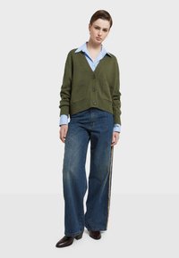 Olivově zelený cardigan s V-výstřihem, třemi knoflíky a dvěma předními kapsami, nošený přes světle modrou pruhovanou košili a kalhoty s širokými nohavicemi.