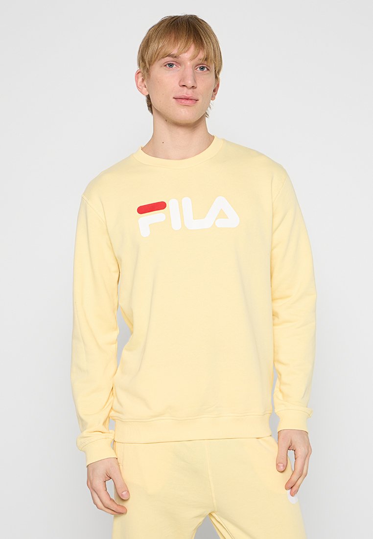 Joven vistiendo un sudadera FILA de color amarillo claro y pantalones a juego, de pie frente a un fondo blanco liso.