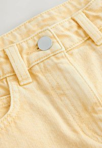 Nærbillede af livvidden på lysegule cowboybukser i denim med sølvknap, bæltestropper og synlige syningsdetaljer.