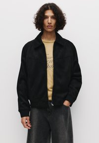 Giacca bomber nera in suede con colletto e zip frontale, abbinata a una t-shirt grafica gialla e pantaloni in denim grigi.