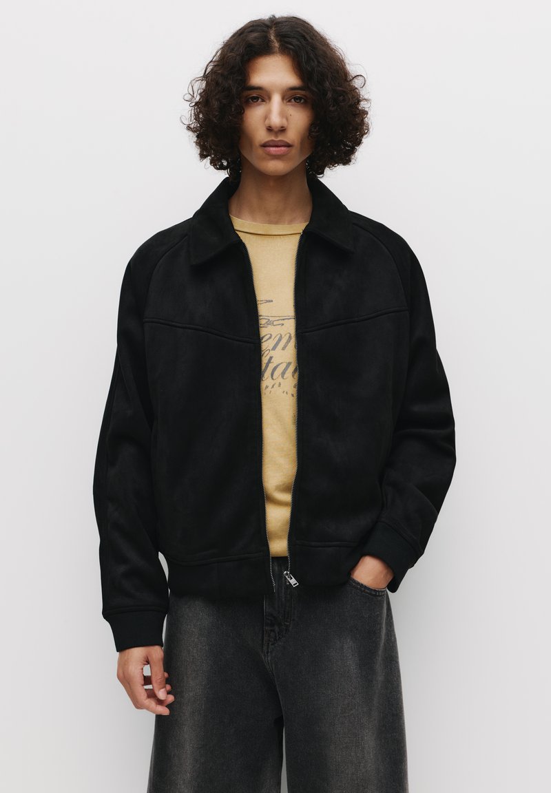 Giacca bomber nera in suede con colletto e zip frontale, abbinata a una t-shirt grafica gialla e pantaloni in denim grigi.