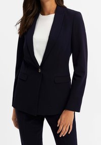 Navy blazer made of smooth fabric, featuring a notched lapel, single button closure, and two front pockets. Styled with a white shirt underneath.