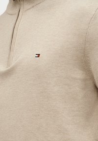 Μπεζ πλεκτό πουλόβερ με φερμουάρ στο ένα τέταρτο και μικρό κεντητό λογότυπο σε κόκκινο, λευκό και μαύρο στο στήθος.