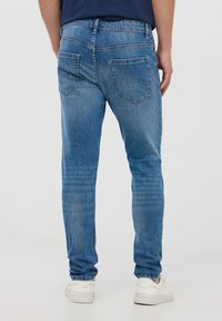 Jeans in denim blu con una forma aderente, caratterizzati da tasche posteriori, un leggero effetto sbiadito e sneaker bianche visibili alla caviglia.