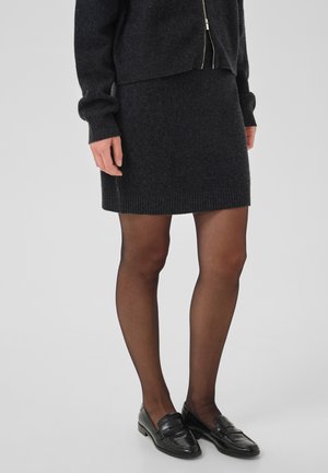 Personne portant un pull foncé zippé, une jupe foncée assortie, des collants noirs transparents et des mocassins noirs, debout devant un fond uni.