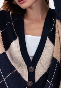 Cardigan à motif losanges bleu marine et beige avec un col en V profond, des boutons et un tissu en tricot texturé. Rehaussé de bordures côtelées foncées.