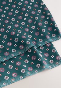 Teal fabric featuring a repeating pattern of circular motifs and flowers in white, red, and cream. Edges have a fringed finish.