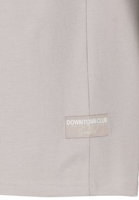 Light gray cotton T-shirt featuring a rectangular brand label with white text sewn on the lower side and a smooth, soft texture.