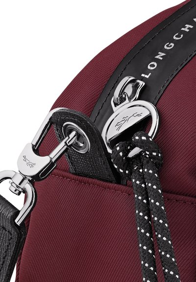 Nahaufnahme des silbernen Verschlusses und des schwarzen Gurtverbinders einer kastanienroten Tasche mit reflektierenden schwarz-weißen Kordeln auf strukturiertem Stoff.