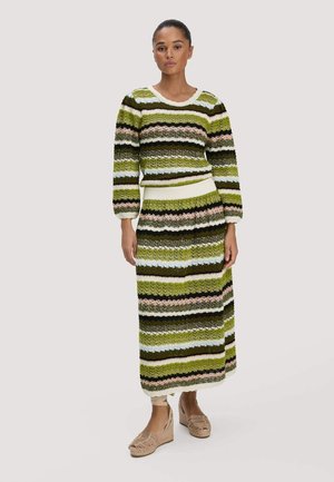 Frau trägt einen gestrickten Pullover mit grünen, braunen, blauen, rosa und weißen Streifen sowie einen passenden langen Rock und beige Keilsandalen.