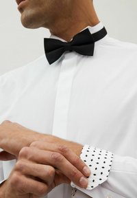 Fekete texturált csokornyakkendő fehér inggel, amelyen fekete, ismétlődő gyémántmintázat található az ujjakon. Zártközeli kép egy kézről, amely az ujját állítja.
