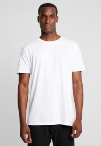 White cotton t-shirt with short sleeves, a crew neck, and a relaxed fit. Smooth texture without patterns. Worn over black pants.