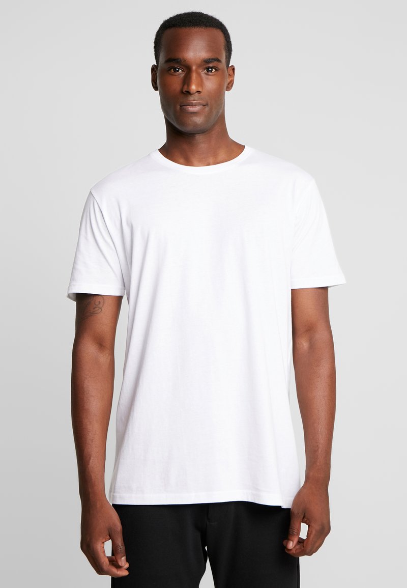 White cotton t-shirt with short sleeves, a crew neck, and a relaxed fit. Smooth texture without patterns. Worn over black pants.