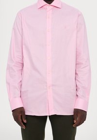 Man som bär en rosa rutmönstrad skjorta med långa ärmar och mörka byxor, stående med armarna avslappnat vid sidorna.