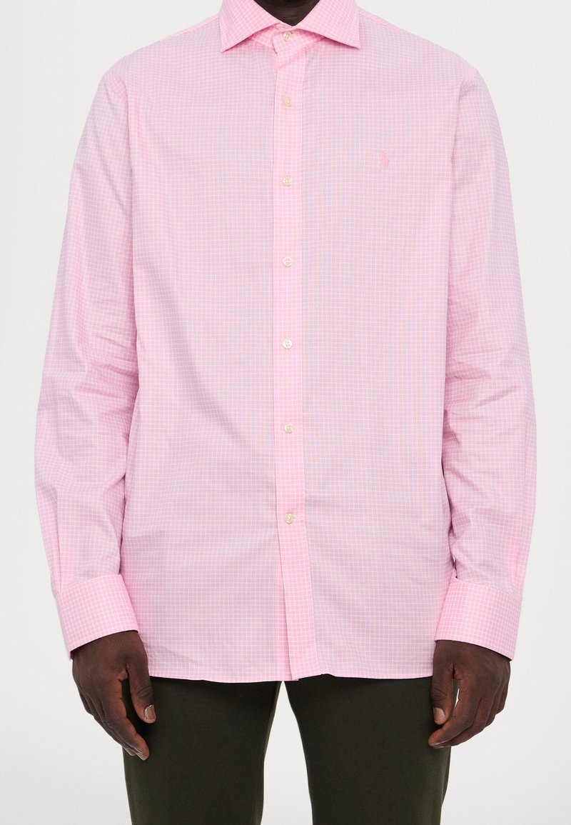 Man som bär en rosa rutmönstrad skjorta med långa ärmar och mörka byxor, stående med armarna avslappnat vid sidorna.