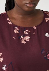 Bordeaux kleurige stof top met een ronde halslijn en een bloemenpatroon in zachte roze en donkerblauwe tinten. Zachte textuur met een lichte glans.