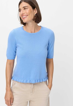Donna sorridente, indossa un maglione azzurro chiaro a maniche corte con orlo arricciato e pantaloni beige con le mani nelle tasche.