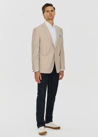 Beżowy lniany blazer z klapami, z dwoma przednimi kieszeniami, zestawiony z białą koszulą, ciemnymi dżinsami i jasnymi sneakersami na gumowej podeszwie.
