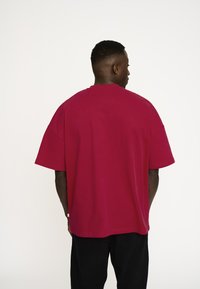 Bordeaux oversized t-shirt i bomuld, med korte ærmer og rund halsudskæring, set bagfra mod en ensfarvet hvid baggrund.