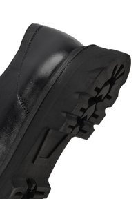 Zapato de cuero negro con un acabado brillante, que cuenta con una suela de goma gruesa con una superficie texturizada y un patrón para mejorar la tracción.