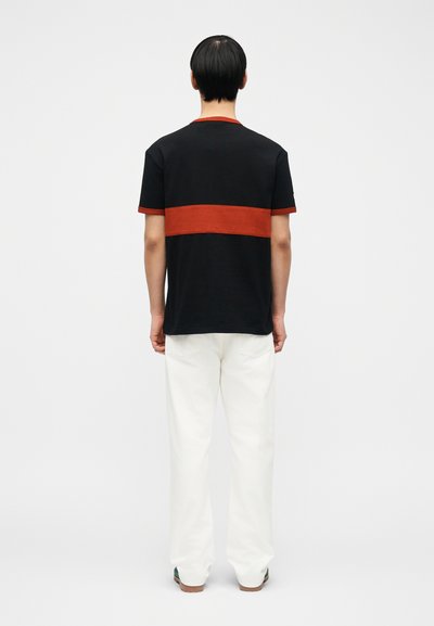 Άνδρας που στέκεται με την πλάτη γυρισμένη, φοράει μαύρο T-shirt με πλατιά κόκκινη οριζόντια ρίγα, άσπρο παντελόνι και σκούρα παπούτσια, σε απλό φόντο.