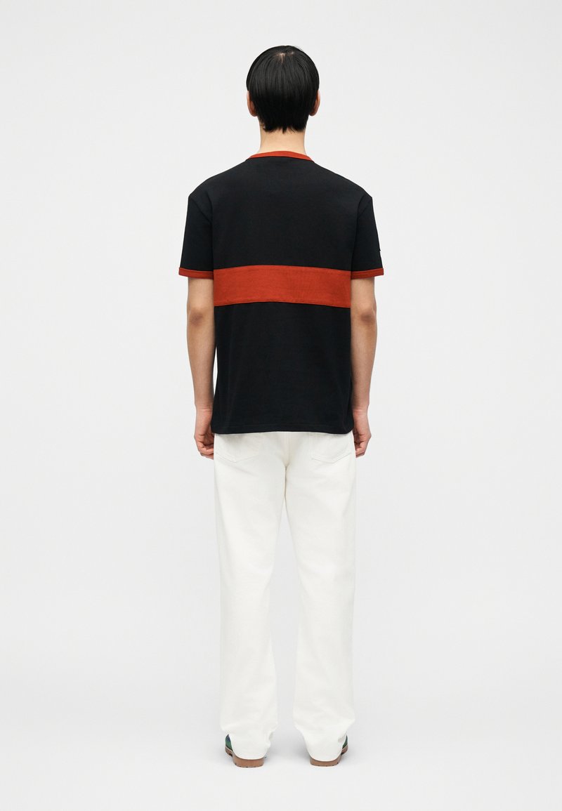 Homme debout, de dos, portant un t-shirt noir avec une large bande horizontale rouge, un pantalon blanc et des chaussures foncées sur un fond uni.