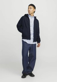 Veste à capuche navy sur une chemise rayée bleu clair, associée à un jean bleu ample et des chaussures noires. La veste a des poches latérales.