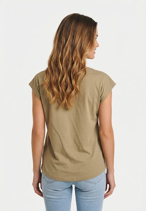 Vrouw met lang golvend bruin haar, gekleed in een olijfgroen shirt met korte mouwen en lichtblauwe spijkerbroek, van achteren gezien tegen een effen achtergrond.