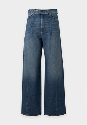 Junge Frau steht mit Händen in den Taschen, trägt ein schwarzes Kurzarm-Rippenoberteil und weite blaue Jeans vor weißem Hintergrund.