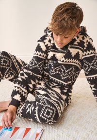 Tuta intera in pile con motivo nero e crema con cappuccio; presenta disegni geometrici. Persona seduta su un tappeto testurizzato, che gioca con dei pezzi di gioco.