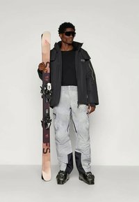 Homme portant une veste de ski noire à capuche, un t-shirt noir, un pantalon de ski gris avec des accents bleus, tenant un ski avec des fixations.