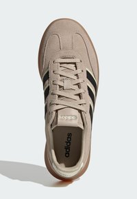 Bež nubuk tenisice s crnim i svijetlozelenim prugama, zaobljen nos, vezanje sprijeda i gumeni potplat. Adidas logo na jeziku i sa strane.