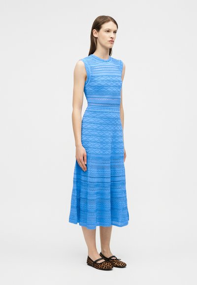 Jeune femme debout portant une robe midi bleu sans manches à motifs et des ballerines léopard, sur un fond clair uni.