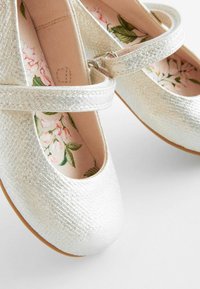 Silverfärgade ballerinas med texturerad yta, justerbar rem och blommönstrad innersula. Rund tå och mjukt foder.