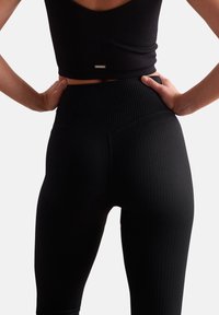 Leggings negras con textura acanalada y una cinturilla alta, que presentan un sutil detalle de logo y una superficie suave, mostrados desde la parte trasera.