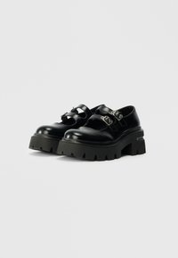Sapatos de plataforma pretos em couro, com duas fivelas ajustáveis, biqueira arredondada e sola grossa em borracha preta texturizada com ranhuras profundas.