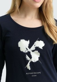Marineblaues Langarmshirt aus weichem Stoff mit einem weißen abstrakten Design und sich kreuzenden Linien sowie dem Text "ALPINE ESCAPE" am unteren Rand.