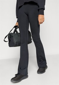 Μαύρα κολάν με καμπάνα και ομαλή υφή, συνδυασμένα με μαύρη τσάντα duffel και μαύρα αθλητικά παπούτσια από δίχτυ, παρουσιασμένα σε ουδέτερο φόντο.