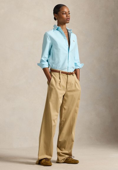 Frau steht mit den Händen in den Taschen, trägt ein hellblaues Hemd mit Knöpfen, beige, weit geschnittene Hose, braunen Gürtel und Mokassin-Schuhe.