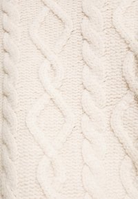 Tissu tricoté de couleur crème avec des motifs torsadés en relief créant des lignes verticales texturées.