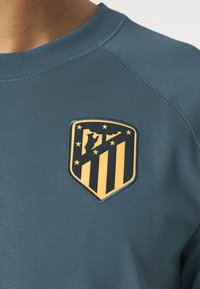 Sudadera azul con el escudo del club de fútbol Atlético de Madrid en amarillo y negro en el pecho cerca del cuello.