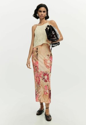 Žena nosi béžový pletený nátelník, midi sukňu s kvetinovým vzorom v ružových a hnedých tónoch, čierne šnurovacie balerínky, drží čiernu tašku pod pažou.