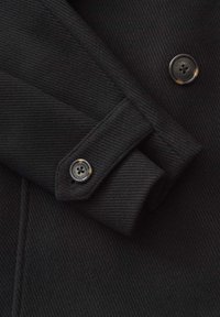 Primo piano di un cappotto di lana nera con un tessuto strutturato, polsino piegato e un pulsante rotondo inciso con un design a quattro fori.